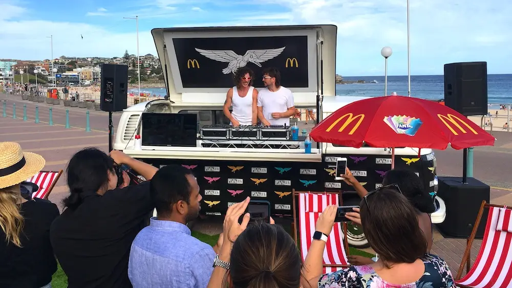 Outdoor brand activation setup with DJ booth and crowd gathered near a branded van by the beach