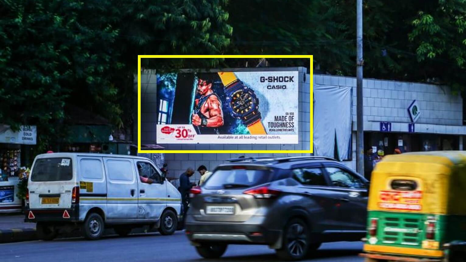 A street-level view of a G-Shock watch advertisement featuring an athletic man. An auto-rickshaw and a silver SUV blur past the sign in traffic.
