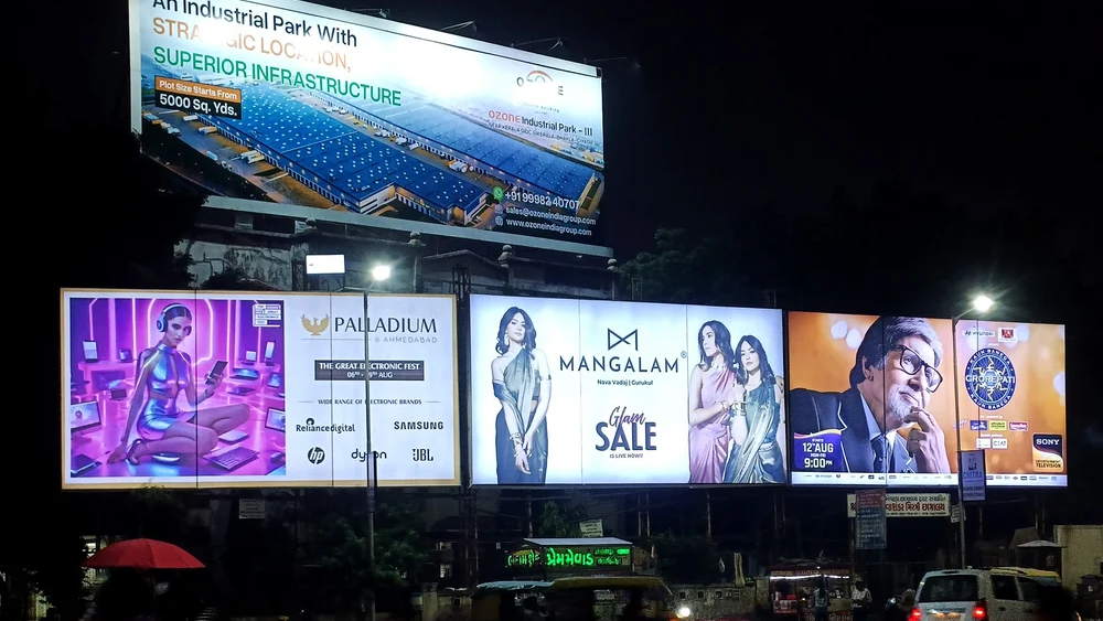 Multiple bright billboards at night showcasing diverse ads, including fashion and electronics, positioned above a busy urban street with passing auto-rickshaws and dark silhouettes.