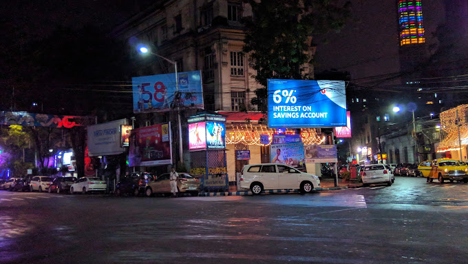 City intersection at night with glowing blue bank advertisements for savings accounts. Parked cars line the street near traditional architecture and a colorful skyscraper.
