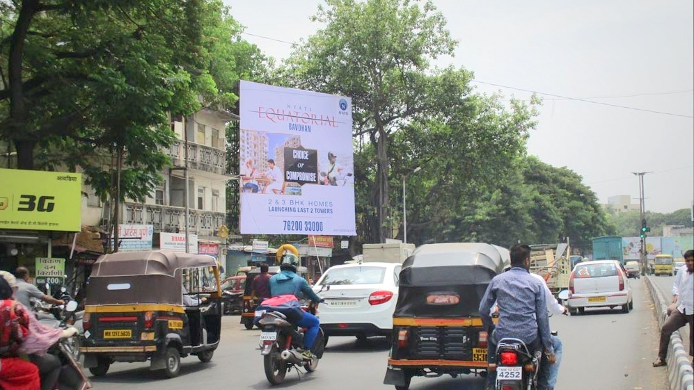 A large vertical billboard for Nyati Equatorial homes stands tall beside a busy street filled with auto-rickshaws, motorcycles, and lush green trees during daytime.