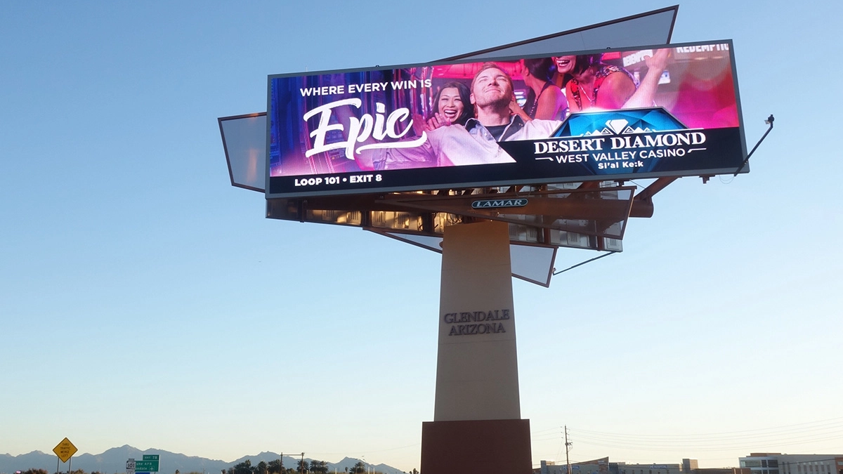 A large digital billboard displays a vibrant "Desert Diamond Casino" advertisement against a clear blue sky. It features the text "Where Every Win Is Epic" alongside joyful people celebrating a win.