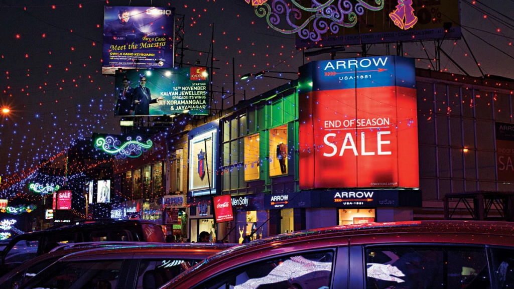 A bustling night scene in an Indian city shows multiple illuminated billboards and neon signs, including a large "End of Season Sale" LED screen for Arrow USA 1851.