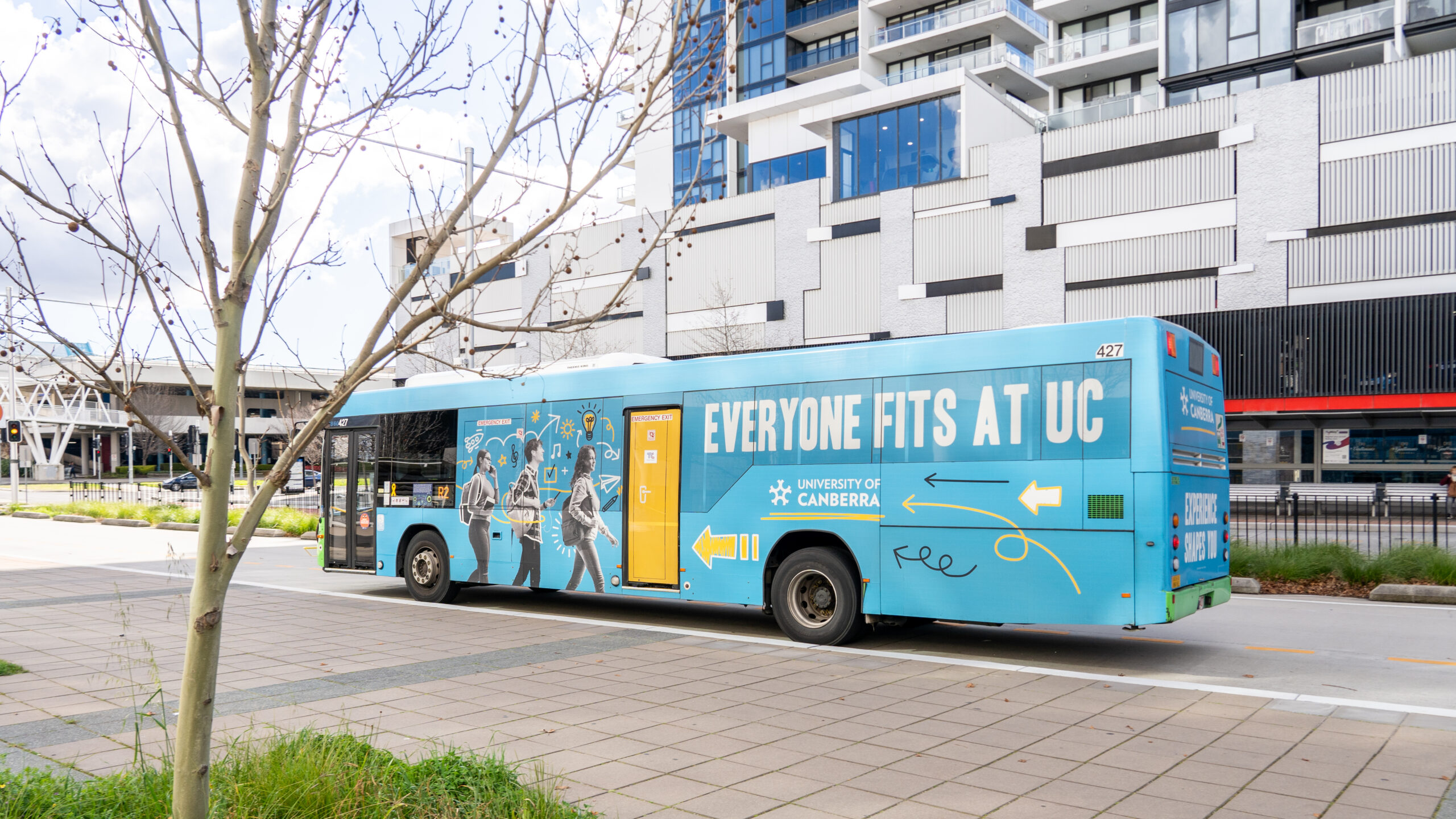A bright blue city bus is fully wrapped with a University of Canberra advertisement featuring the slogan "Everyone Fits at UC" alongside illustrations of students and academic icons.