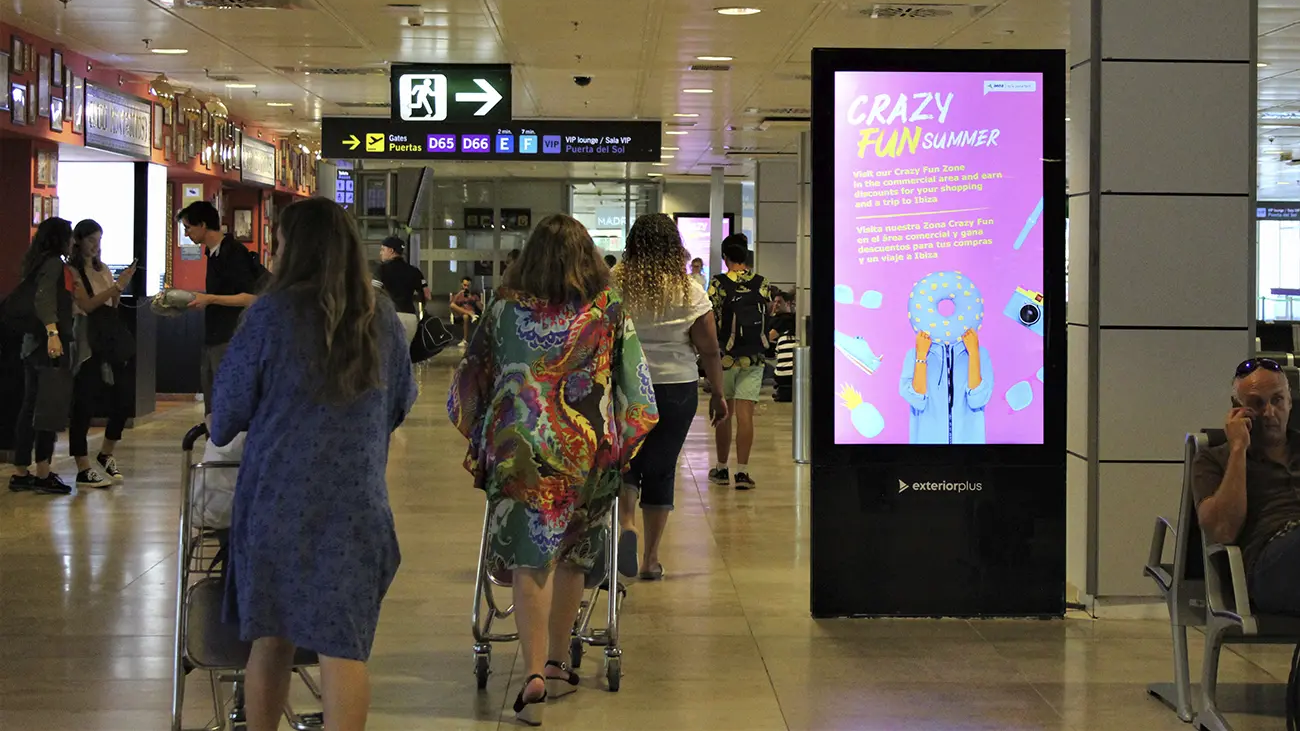 A tall digital kiosk in a busy airport terminal displays a bright purple "Crazy Fun Summer" advertisement featuring a person holding a donut, positioned amidst traveling passengers and luggage.