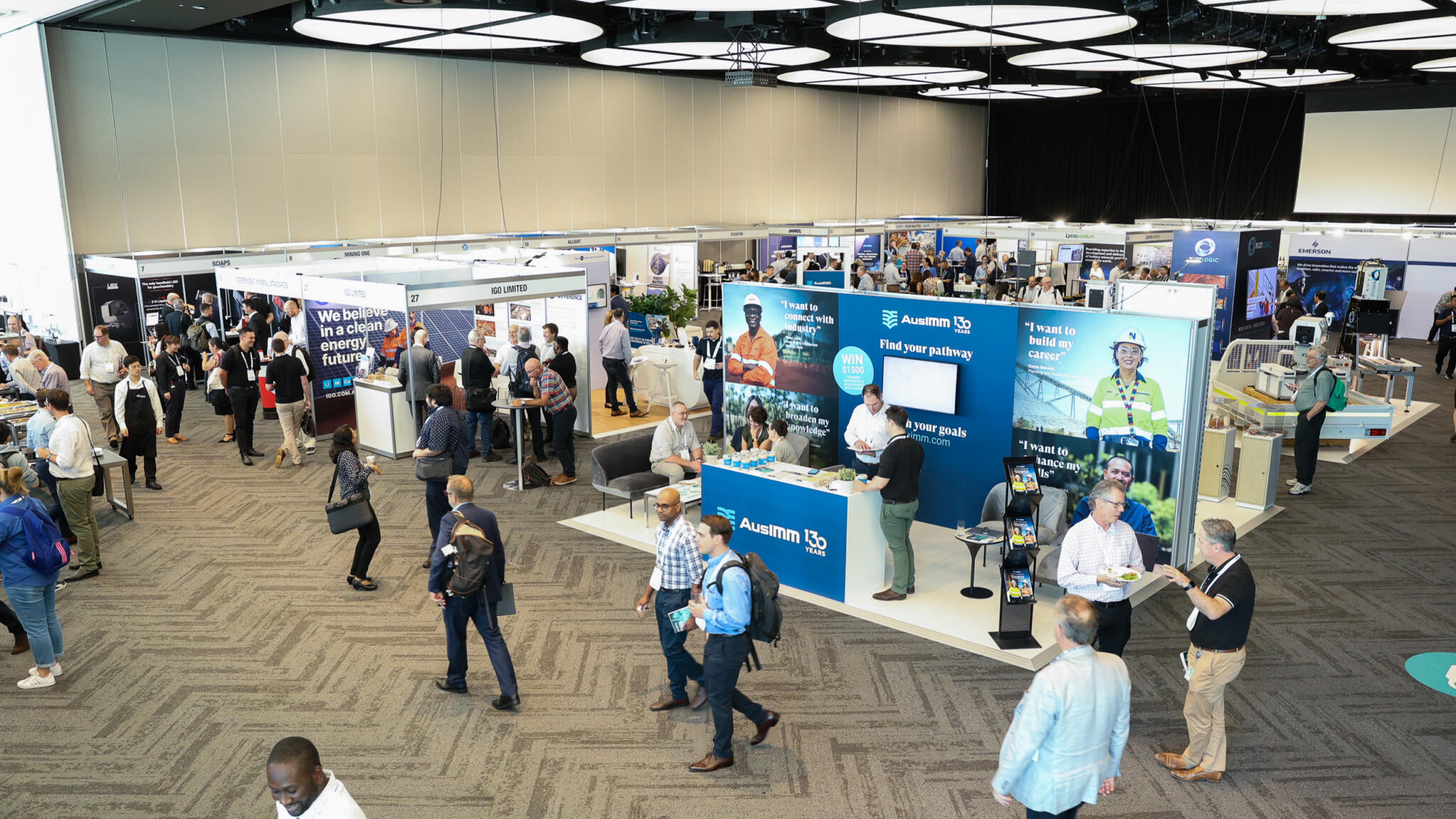 An expansive indoor convention hall filled with professional trade show booths, featuring large-scale printed graphics, digital displays, and branding for the AusIMM 130-year anniversary and various mining companies.