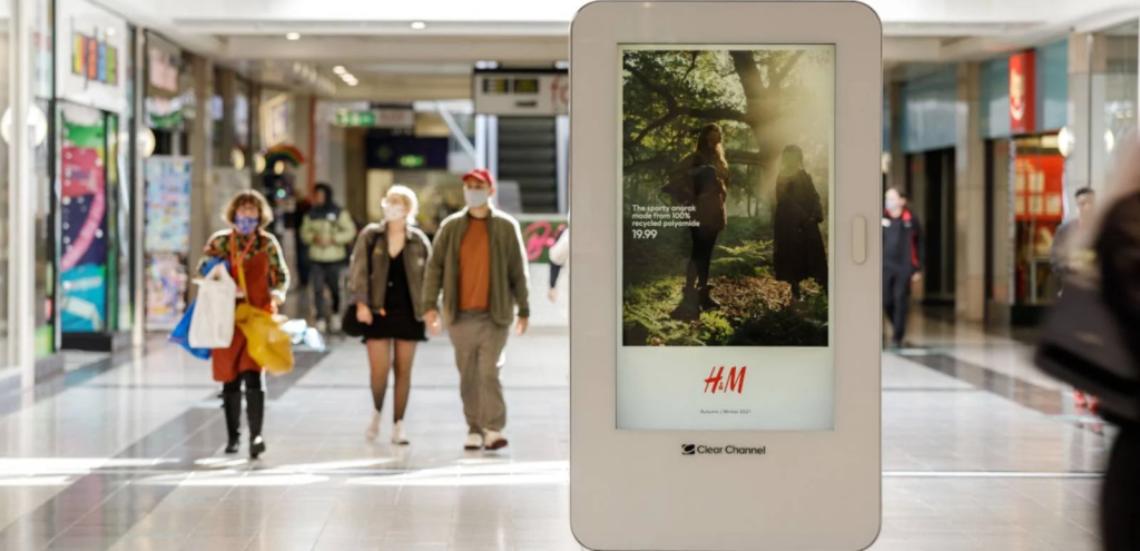 A sleek, white digital pedestal stands in a brightly lit shopping mall corridor, displaying a high-fashion H&M "Autumn/Winter" advertisement to shoppers walking through the gallery.