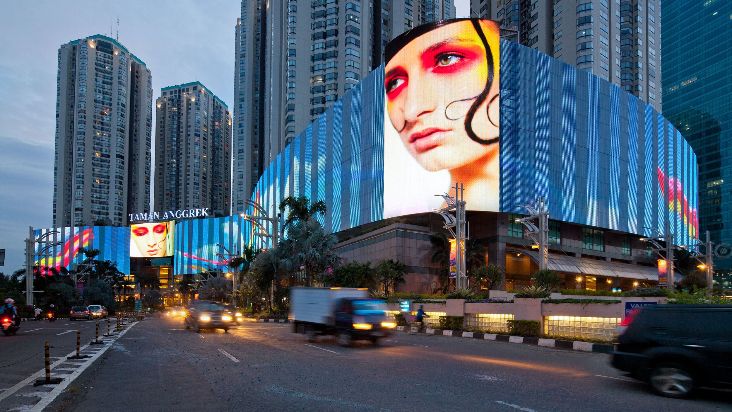A vibrant, massive curved LED facade on the Taman Anggrek building displays a high-fashion close-up portrait of a woman with red eye makeup, towering over a busy city street at dusk.