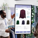 Retail Interactive Kiosk: A store associate demonstrates a large touch-screen vertical kiosk that allows customers to browse apparel, select colors/sizes, and make purchases directly from the shop floor.