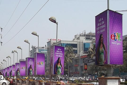 A series of illuminated median pole kiosks display eBay advertisements along a busy urban roadway. Positioned at regular intervals, the lamp post advertising installation creates repeated brand exposure for motorists and commuters, turning the entire corridor into a high visibility outdoor media zone.