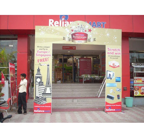 A promotional arch gate installed at the entrance of a Reliance Mart store to celebrate its anniversary campaign. The branded entry structure features festive graphics, prize promotion messaging, travel giveaway visuals, and scratch-and-win offers on both side pillars. Positioned directly at the store entrance, the arch gate transforms the retail frontage into a high-visibility promotional touchpoint that attracts shoppers and drives campaign engagement.