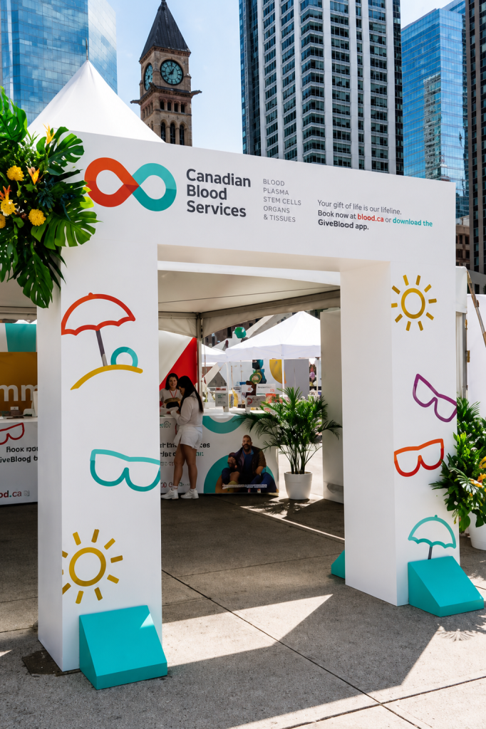A large custom branded arch gate for Canadian Blood Services installed at an outdoor public event, featuring clean white panels, colorful summer themed graphics, and campaign messaging. Positioned at the entrance of the activation space, the archway creates a welcoming brand touchpoint while guiding visitors into the experiential engagement area surrounded by event booths and city skyline views.