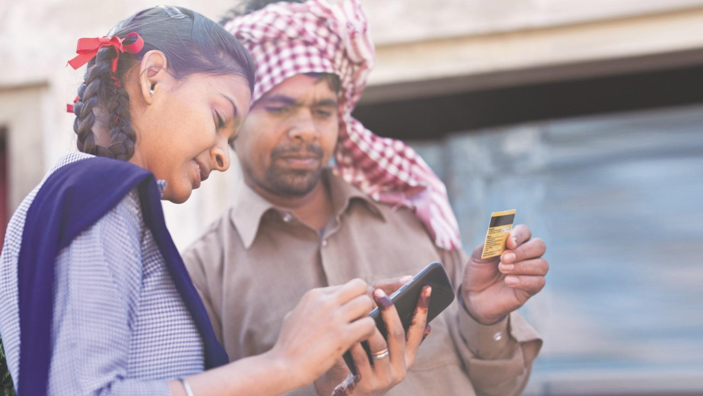 A rural payment interaction showing a young schoolgirl using a smartphone while a man beside her holds a bank card, highlighting digital payment adoption and mobile banking access in a village setting.