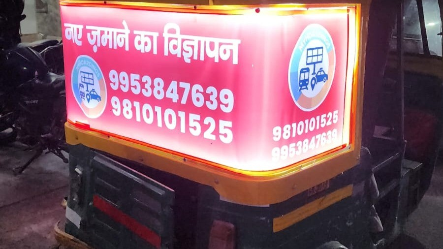 An illuminated auto-rickshaw back panel advertisement featuring Hindi text, contact numbers, and transport-themed icons, showcasing a local mobile advertising format commonly used for regional outdoor promotions in India.