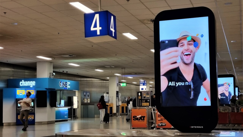 A digital airport advertising display installed inside an airport terminal, featuring a vertical video-style screen showing a mobile app promotion with a person taking a selfie. The high-visibility airport advertisement is placed near passenger walkways and waiting areas to capture traveler attention in a busy transit environment.