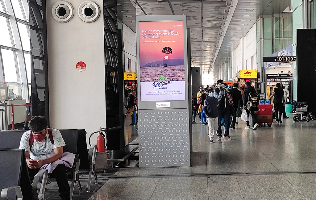 A digital airport advertising display placed inside a busy airport terminal, featuring a tourism and lifestyle campaign promoting an event in Hirakud, Odisha. The illuminated vertical screen is positioned along a passenger walkway to attract traveler attention in a high-footfall airport environment.