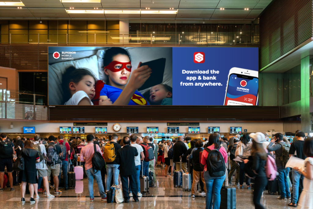 A large digital airport advertising billboard displayed above a crowded airport check-in area, featuring a mobile banking app campaign with a superhero-themed visual and smartphone graphic. The high-impact airport advertisement is positioned to capture the attention of travelers and passengers in a busy terminal environment.