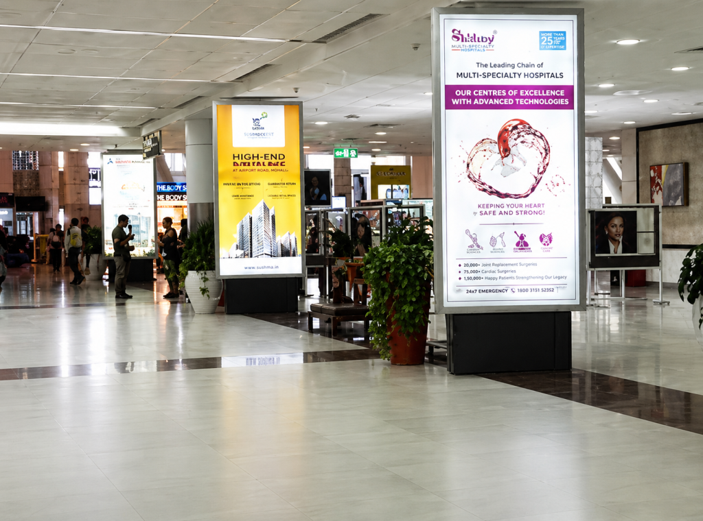 A series of illuminated airport advertising displays installed inside a busy airport terminal, featuring healthcare and real estate promotional campaigns on large vertical digital panels. The high-visibility airport advertisements are positioned along passenger walkways to capture traveler attention in a premium transit environment.