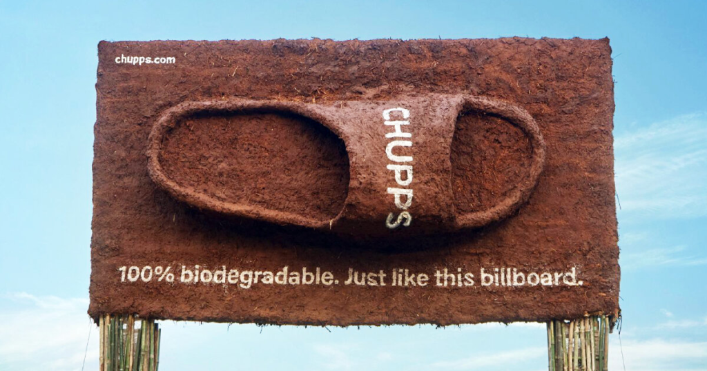 A creative eco-friendly billboard made entirely from biodegradable natural materials displays a large slipper-shaped product with the message “100% biodegradable. Just like this billboard.” The sustainable outdoor advertising campaign highlights environmentally conscious branding through biodegradable billboard construction and earthy visual design.
