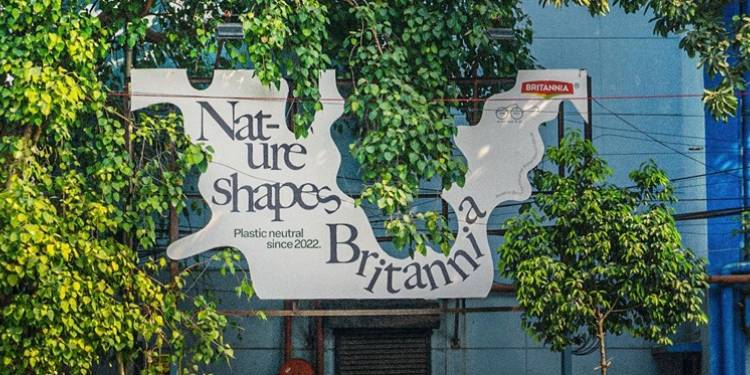 A creatively shaped Britannia billboard surrounded by lush green trees promotes sustainability with the message “Nature Shapes Britannia” and “Plastic neutral since 2022.” The eco-conscious outdoor advertising campaign blends organic billboard design with natural surroundings to emphasize environmental responsibility and sustainable branding.