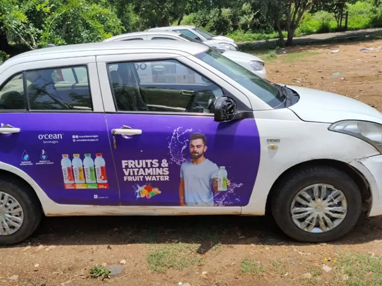 A branded cab advertising campaign features a car wrap promoting a fruit water beverage brand with vibrant purple graphics, product visuals, and celebrity endorsement imagery displayed across the vehicle doors. The transit advertising setup uses mobile cab branding and on-road outdoor advertising to increase brand awareness and visibility in high-traffic urban areas.