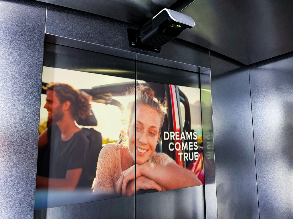 An elevator branding campaign inside a modern lift features a large lifestyle advertisement displayed across the elevator doors with the message “Dreams Comes True.” The indoor advertising setup uses elevator door branding and immersive visual marketing to engage passengers in high-traffic commercial and residential spaces.