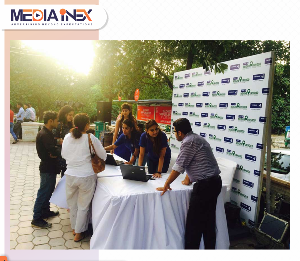 A society activation event is taking place in an outdoor residential community space where brand representatives are interacting with residents at a registration and engagement desk. The setup includes a branded backdrop, information counter, and on-ground staff assisting visitors, creating direct community outreach, consumer interaction, and localized experiential marketing within the neighborhood environment.