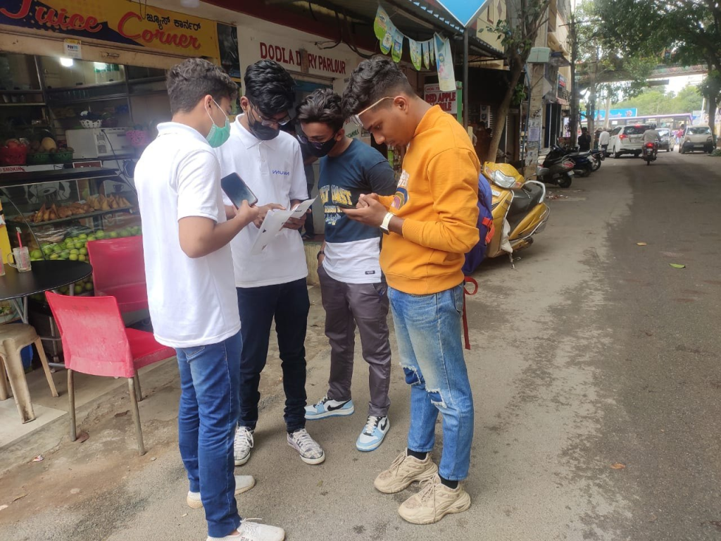 A group of young brand promoters is conducting a street-level on-ground activation campaign near a local market area, engaging directly with pedestrians through surveys, registrations, or promotional interactions using mobile devices and printed materials. The activity reflects direct consumer outreach and face-to-face engagement in a real urban environment.