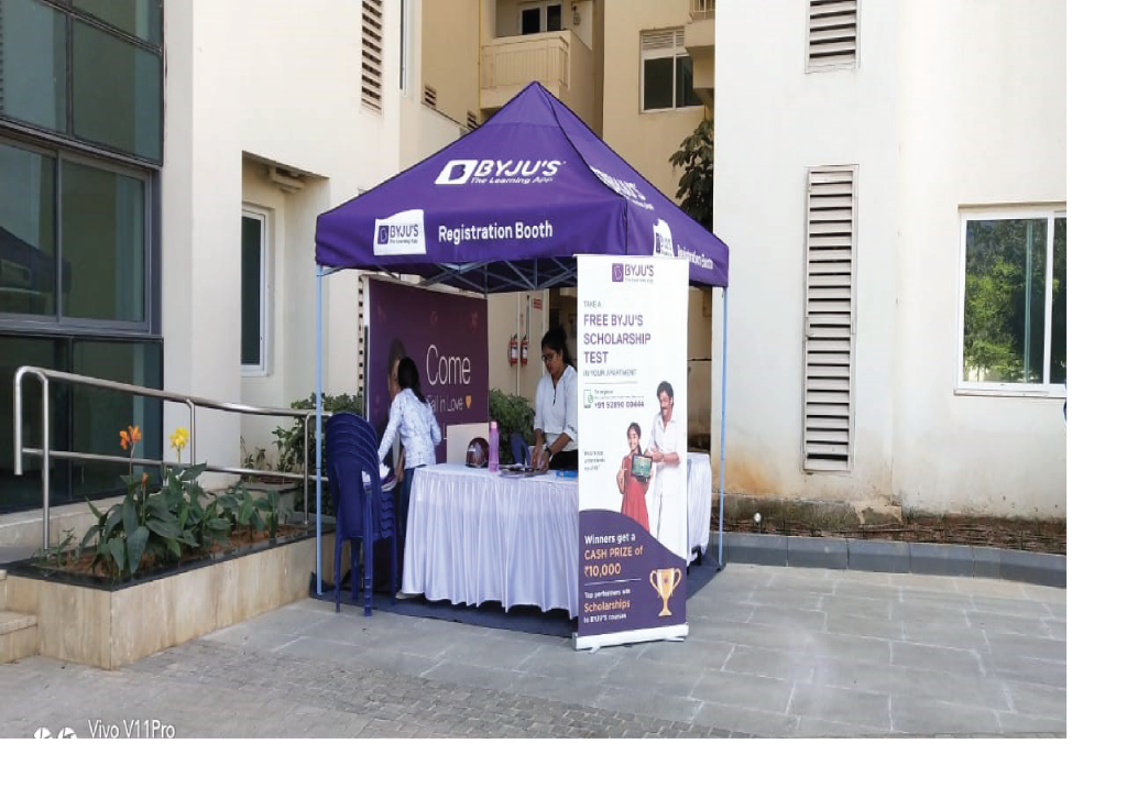 A BYJU’S society activation setup is installed inside a residential apartment complex featuring a branded registration booth for a scholarship test campaign. The on-ground activation includes promotional standees, a help desk, and brand representatives engaging directly with residents and parents to encourage participation, creating localized educational outreach and community engagement.