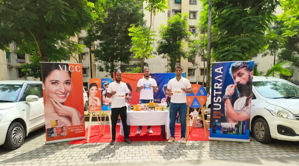 A society activation campaign featuring VLCC and Ustraa is set up inside a residential community area with branded standees, product displays, and promotional counters. Brand representatives are engaging residents through product sampling and interactive on-ground marketing activities, creating direct consumer interaction and lifestyle-focused brand awareness within the society environment.