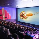 Cinema advertising displayed on a large movie theatre screen featuring a food brand promotion, capturing the attention of a seated audience in a packed cinema hall.