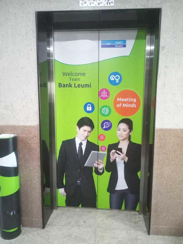 Elevator door branding campaign featuring a full-wrap advertisement for Bank Leumi, displaying business professionals, corporate messaging, and brand visuals across the elevator doors in an office building.