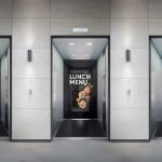 Elevator interior branding displaying full-height digital advertisements on the back walls of multiple elevators, showcasing fashion, restaurant, and promotional campaigns in a modern commercial building.